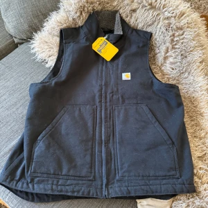 Svart väst från Carhartt - Säljer en svart väst från Carhartt med dragkedja och två stora fickor framtill. Västen har en hög krage och är fodrad med sherpa för extra värme. Perfekt för kyligare dagar. Helt oanvänd, säljer för att den var för stor. Nypris 1700kr!