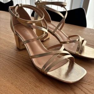 Guldiga sandaler med klack - Snygga guldiga sandaler med klack och remmar som korsar över foten. De har en fyrkantig tå och justerbart spänne vid ankeln. Endast använda vid ett tillfälle, som nya! Perfekta för fin tillställning som bal💕 6cm klack