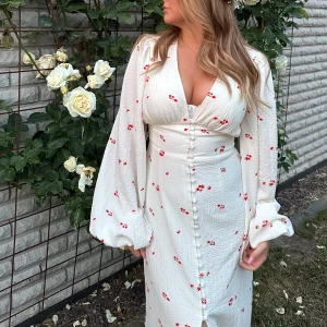 Positano Dress- Adoore - Hej, jag säljer ju min Adoore klänning då den inte kommer till användning. Endast använd en gång. En charmig vit klänning med små röda blommor och knappar framtill. Den har långa puffärmar och en v-ringad design som ger en romantisk känsla. Perfekt nu till sommaren 🌸