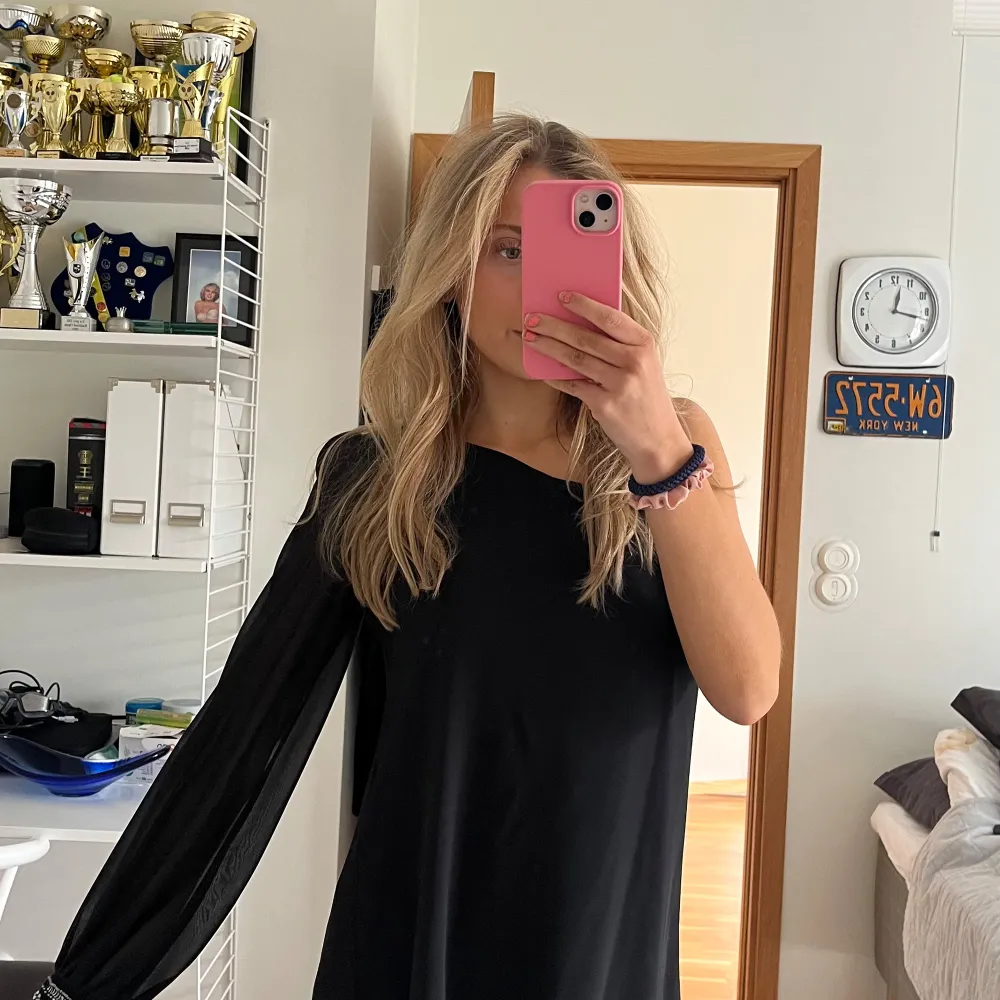 Snygg svart oneshoulder klänning med lång ärm och dekorativa pärlor vid ärmslutet. Perfekt för en stilren look.. Mekot.