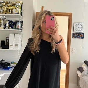 Svart oneshoulder klänning - Snygg svart oneshoulder klänning med lång ärm och dekorativa pärlor vid ärmslutet. Perfekt för en stilren look.