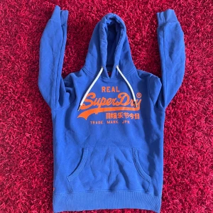 Blå Super Dry Hoodie - Säljer en blå hoodie från Superdry med orange tryck på framsidan. Tröjan är i mycket bra skick. Storlek M passar lite större. Skriv vid minsta fundering