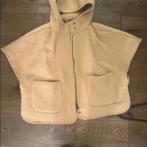 Beige väst med huva - Snygg beige väst med huva och dragkedja framtill. Västen har två stora fickor och knappar vid kragen. Perfekt för lager-på-lager-stil.