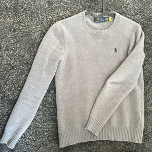 Grå tröja från Polo Ralph Lauren - Säljer en stilren grå tröja från Polo Ralph Lauren med deras ikoniska logga broderad på bröstet. Tröjan har långa ärmar och ribbade muddar vid ärmslut och nederkant. Perfekt för en klassisk och avslappnad look. Är mycket noppig