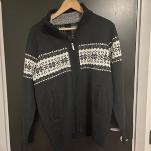 Stickad kofta - Säljer en svart stickad kofta med ett vitt mönster över bröstet och ärmarna. Koftan har dragkedja och en mysig fodrad insida i rutigt mönster. Perfekt för kyliga dagar! Väldigt fin passform!