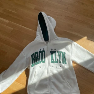 Vit hoodie med Brooklyn-tryck från Shein - Säljer en vit hoodie från Shein med ett coolt Brooklyn-tryck i grönt och svart. Hoodien har dragkedja och en mysig huva med mörkgrönt foder. Perfekt för en avslappnad stil! 🗽