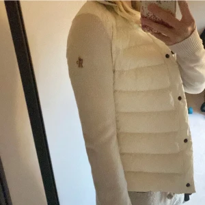 Vit dunväst från Moncler - Säljer en stilren vit dunväst från Moncler med knappar framtill. Västen har en quiltad design och är perfekt för vår dagarna . Den har en broderad logga på ärmen som ger en exklusiv touch.