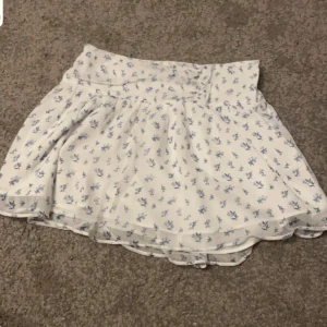 Vit blommig kortkjol - Söt vit kortkjol med blå blommigt mönster. Kjolen har en volangdetalj som ger den en extra touch av stil. Perfekt för varma dagar och kan enkelt matchas med olika toppar.