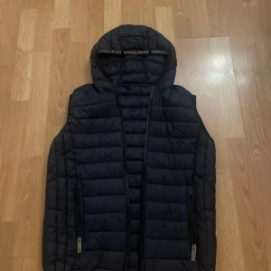 Svart dunväst från Emporio Armani - Snygg svart dunväst från Emporio Armani med huva och dragkedja. Perfekt för kyligare dagar när du vill hålla stilen. Västen har en quiltad design och är både praktisk och trendig.