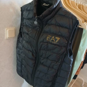 Svart dunväst från Emporio Armani - Snygg svart lättviktsväst från Emporio Armani med dragkedja och märkets logga i guld på bröstet. Västen har en quiltad design och hög krage. Inga skador eller märken. Kan användas av både dam,herr eller ungdom.