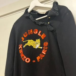 Svart tröja från Kenzo x H&M - Säljer en svart tröja från Kenzo x H&M med ett färgglatt tryck av en tiger och texten 'Jungle Kenzo - Paris'. Tröjan har volangdetaljer vid halsen och knappar med guldfärgade detaljer.