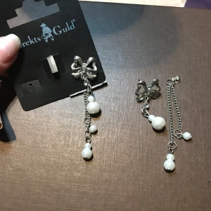 Örhängen med rosett och pärlor från Albrekts Guld - Eleganta örhängen från Albrekts Guld med en vacker rosett i silverfärg och hängande kedjor prydda med vita pärlor. Perfekta för att ge en stilfull touch till din look.