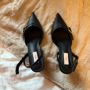 Svarta klackskor med spetsig tå - Snygga svarta klackskor från Nelly med spetsig tå och elegant remdesign. Skorna har en justerbar rem med spänne och små nitar för extra stil. Perfekta för en chic look.