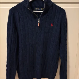 Mörkblå stickad tröja från Polo Ralph Lauren - Säljer en snygg mörkblå stickad tröja från Polo Ralph Lauren med dragkedja och det klassiska röda logotypbroderiet på bröstet. Perfekt för kyliga dagar med sin kabelstickade design och ribbade detaljer vid ärmslut och nederkant.
