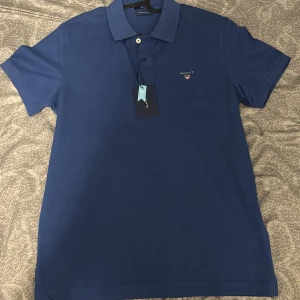 Blå pikétröja från GANT - Säljer en stilren blå pikétröja från GANT. Tröjan har en klassisk krage med knappar och GANT-loggan broderad på bröstet. Perfekt för en avslappnad men ändå snygg look. 💙