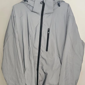 Grå reflex vindjacka från Biltema - Säljer en grå reflex -vindjacka från Biltema i storlek XXL. Jackan har en dragkedja framtill och en praktisk huva. Tillverkad i 100% polyester, perfekt för blåsiga dagar. Jackan är oanvänd. 