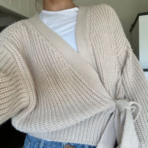 Beige stickad omlottkofta från Zara - Säljer en mysig beige stickad kofta från Zara. Den har en omlott-design med knytband i midjan och långa ärmar. Perfekt för kyliga dagar!