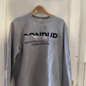 Grå sweatshirt från Dondup - Säljer en grå sweatshirt från Dondup med texten 'Unconventional Clothing Established 1999' tryckt på framsidan. Tröjan har en rund halsringning och långa ärmar. Perfekt för en avslappnad stil.