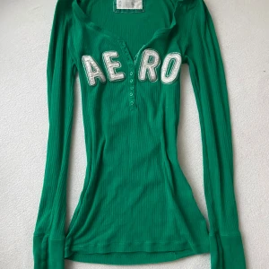 Grön ribbad långärmad topp från Aeropostale - Säljer en grön ribbad långärmad topp från Aeropostale med texten 'AERO' i vita bokstäver på framsidan. Toppen har en knappdetalj vid halsringningen och är perfekt för en avslappnad stil.