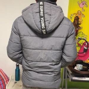 Grå pufferjacka med text på huvan - Säljer en grå pufferjacka med texten 'SUPPLY YOUNG' på huvan. Jackan har en dragkedja framtill och är långärmad. Perfekt för kyligare dagar.
