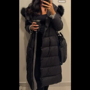 Svart lång dunjacka med pälskrage - Säljer en svart lång dunjacka med pälskrage och huva. Jackan har en stilren design med långa ärmar och är perfekt för kalla dagar. Den är både varm och bekväm, idealisk för vintern.