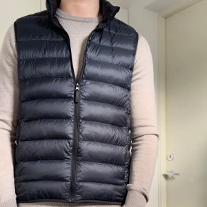 Uniqlo väst - Snygg mörkblå dunväst med dragkedja framtill. Perfekt för lager-på-lager under kyliga dagar. Västen har en quiltad design och hög krage för extra värme.