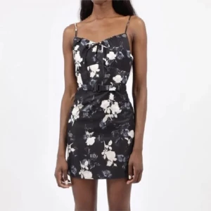 Svart blommig klänning med smala axelband - Oanvänd svart klänning med vita blommor och smala axelband från Wasted Paris. Den har en knytning framtill och ett bälte i midjan för en fin passform. Perfekt för en somrig look! FR S/EU XS