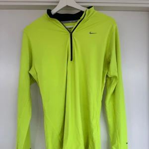 Gul/grön långärmad tröja från Nike - Säljer en gul/grön långärmad tröja från Nike med dragkedja vid halsen. Perfekt för löpning med Dri-FIT-teknologi som håller dig torr och bekväm. Tröjan har en sportig design och är idealisk för träning.