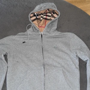 Grå hoodie från Burberry - Säljer en stilren grå hoodie från Burberry med dragkedja och klassiskt rutigt mönster på insidan av huvan. Tröjan har långa ärmar och en justerbar huva med snörning. Perfekt för en avslappnad och trendig look.