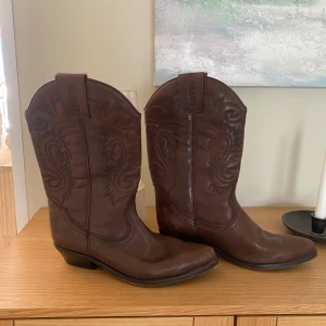 Bruna cowboy boots från Levi's - Snygga bruna cowboy boots från Levi's i skinn med dekorativa broderier. Tillverkade i Spanien, dessa boots har en klassisk design med rund tå och låg klack. Perfekta för en stilren look.