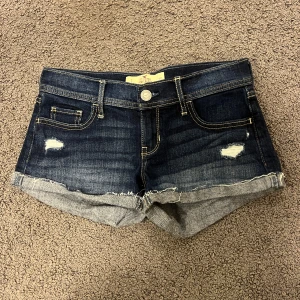Mörkblå lågmidjade jeansshorts från Hollister - Snygga mörkblå jeansshorts från Hollister med slitna detaljer och fransig kant. Perfekta för en avslappnad sommarlook! Middjemåttet på shortsen raktöver är 38 cm och längden är 22 cm och de är ganska strechiga!