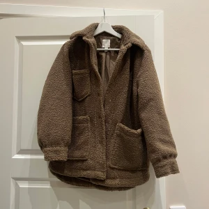 Brun teddyjacka från H&M - Helt oanvänd och jättemysig brun teddyjacka från H&M med långa ärmar och flera praktiska fickor. Perfekt för kyliga dagar när du vill hålla dig varm och bekväm. Jackan har en avslappnad passform och stängs med knappar framtill. ❤️