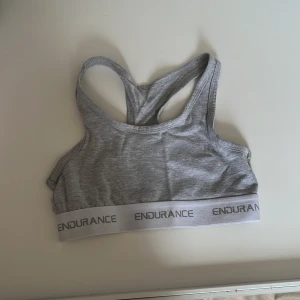 Grå sporttopp från Endurance - Säljer en grå sporttopp från Endurance med brottarrygg och elastiskt band med logotyp. Perfekt för träning och ger bra stöd. Bekväm och stilren design.