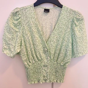 Grön blommig blus från Gina Tricot - Säljer en söt grön blommig blus från Gina Tricot. Blusen har korta ärmar och en v-ringad design med knappar framtill. Den elastiska midjan ger en snygg passform. Perfekt för vår och sommar! 