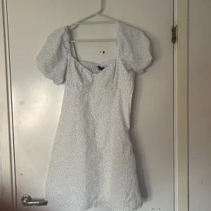 Vit blommig klänning från Divided - Söt vit klänning från Divided med små blå blommor. Den har puffiga kortärmar och en smockad rygg för extra komfort. Perfekt för en somrig look! Den är använd en gång 🪻