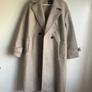Beige kappa från Zara - NY - Säljer en stilren beige kappa från Zara med dubbelknäppning och stora slag. Kappan har långa ärmar med dekorativa knappar vid ärmsluten och praktiska fickor framtill. Perfekt för en elegant look!