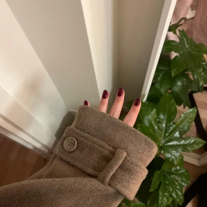 Beige kappa med knappar - Säljer en stilren beige kappa med knappar och hög krage. Jackan har långa ärmar och en klassisk design som passar perfekt för höst och vår. Den är tillverkad i ett mjukt material som ger en bekväm passform.