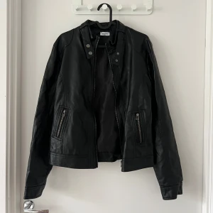 Svart skinnjacka från Pimkie - Snygg svart skinnjacka från Pimkie med dragkedja och knappar. Jackan har en cool bikerstil med två dragkedjefickor framtill och detaljer vid kragen. Perfekt för att ge din outfit en edgy touch. Se baksida för skavank. Inget som man stör sig på. 