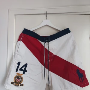 Ralph Lauren vita och röda shorts - Snygga vita shorts från Ralph Lauren med bred röd diagonal rand, mörkblå midja och snörning framtill. Stora broderade detaljer med nummer 14 och Polo-logga. Perfekta för sommaren och strandhäng.