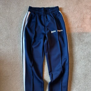 Palm Angels trackpants - Snygga marinblå trackpants från Palm Angels med vita sidoränder och logga på låret. Byxorna har elastisk midja, raka ben och dragkedjor vid bensluten. (Replika)