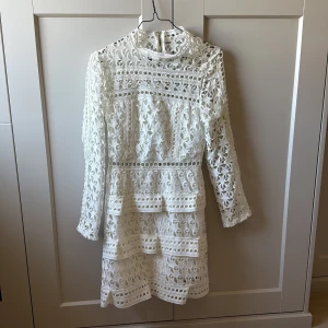 Vit spetsklänning från Borninstockholm - Superfin vit kortklänning från Borninstockholm med långärmade ärmar och vackra spetsdetaljer. Klänningen har hög hals, dragkedja bak och olika detaljer på. 