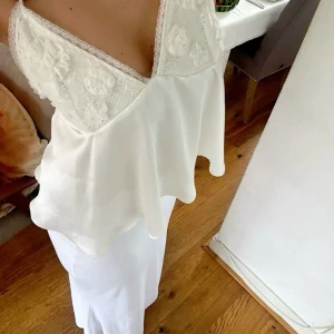 Vit spetsdetaljerad satin topp - Supersöt vit linnetopp med tunna axelband och v-ringning. Toppen har spets med blommiga broderier över bysten och en flowy, lätt satinliknande känsla i tyget. Knytning i ryggen för extra snygg passform. Perfekt för sommarens alla tillfällen!