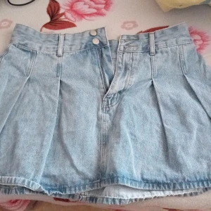 Ljusblå jeanskjol med hög midja - Snygg ljusblå jeanskjol med hög midja och två knappar framtill. Kjolen har bred linning, bälteshällor och veckade detaljer som ger en cool och avslappnad look. Perfekt till sneakers eller boots.