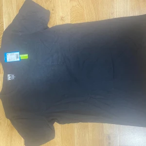 Svart t-shirt med bröstficka från WA - En stilren svart t-shirt från WA i 100% bomull. T-shirten har klassisk passform, rund halsringning och en enkel bröstficka. Perfekt basplagg för dig som gillar en clean look. Gjord av ekologisk bomull för extra komfort.