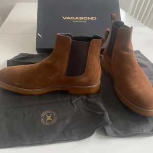 Bruna chelsea boots i mocka från Vagabond - Snygga bruna chelsea boots från Vagabond i mjuk mocka. Skorna har klassisk rund tå, resår på sidorna och dragflik bak för enkel påtagning. Perfekta för dig som vill ha en stilren och tidlös look med premiumkänsla.