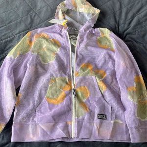 Lila blommig vindjacka från Levi's - Säljer en tunn och luftig vindjacka från Levi's i lila med stora blommor i grönt och orange. Jackan har huva, dragkedja framtill och två stora fickor. Tillverkad i 100% återvunnen polyester, perfekt för dig som vill sticka ut med en färgglad och unik stil.