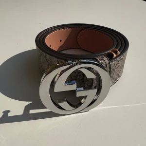 Gucci GG Supreme bälte med silverdetalj - Snyggt bälte från Gucci med klassiskt GG Supreme-mönster i beige och brunt. Bältet har en bred design och en stor, rund spänne i blankt silver med dubbla G-loggan. Tillverkat i skinn med canvas-yttersida och stilren finish. Perfekt för att lyfta vilken outfit som helst.