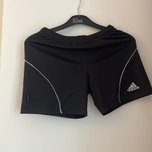 Svarta träningsshorts från Adidas - Svarta träningsshorts från Adidas med vit logga och tunna vita linjer längs sidorna. Shortsen har elastisk midja och är tillverkade i ett lätt och smidigt material som passar perfekt för sport och träning.