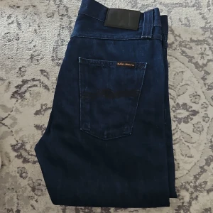 Nudie Jeans Steady Eddie Dry Blue - Säljer ett par mörkblå Steady Eddie jeans från Nudie Jeans i modellen Dry Blue. Jeansen är tillverkade av 100% ekologisk bomull, har klassisk femficksdesign, knappgylf och snygga broderade bakfickor. Perfekta för dig som gillar stilrena och tidlösa jeans.