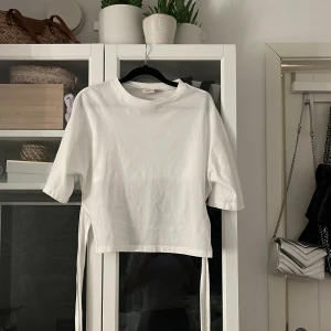 Vit topp från Nelly - Säljer en stilren vit topp från Nelly i storlek XS. Toppen har trekvartsärmar och en unik design med knytband i sidan. Perfekt för en trendig look!